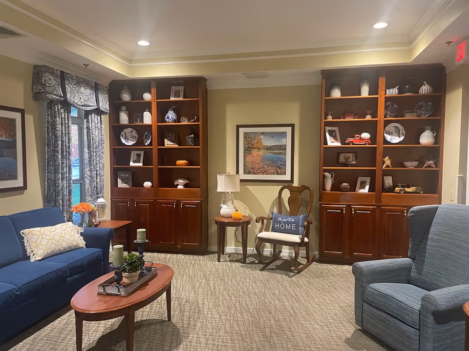 A cozy living room with a blue sofa, a wooden coffee table with candles and a plant, a wooden rocking chair with a cushion that says 'Home', and a blue armchair. The room features built-in wooden shelves filled with decorative items, framed artwork on the walls, and a window with patterned curtains.