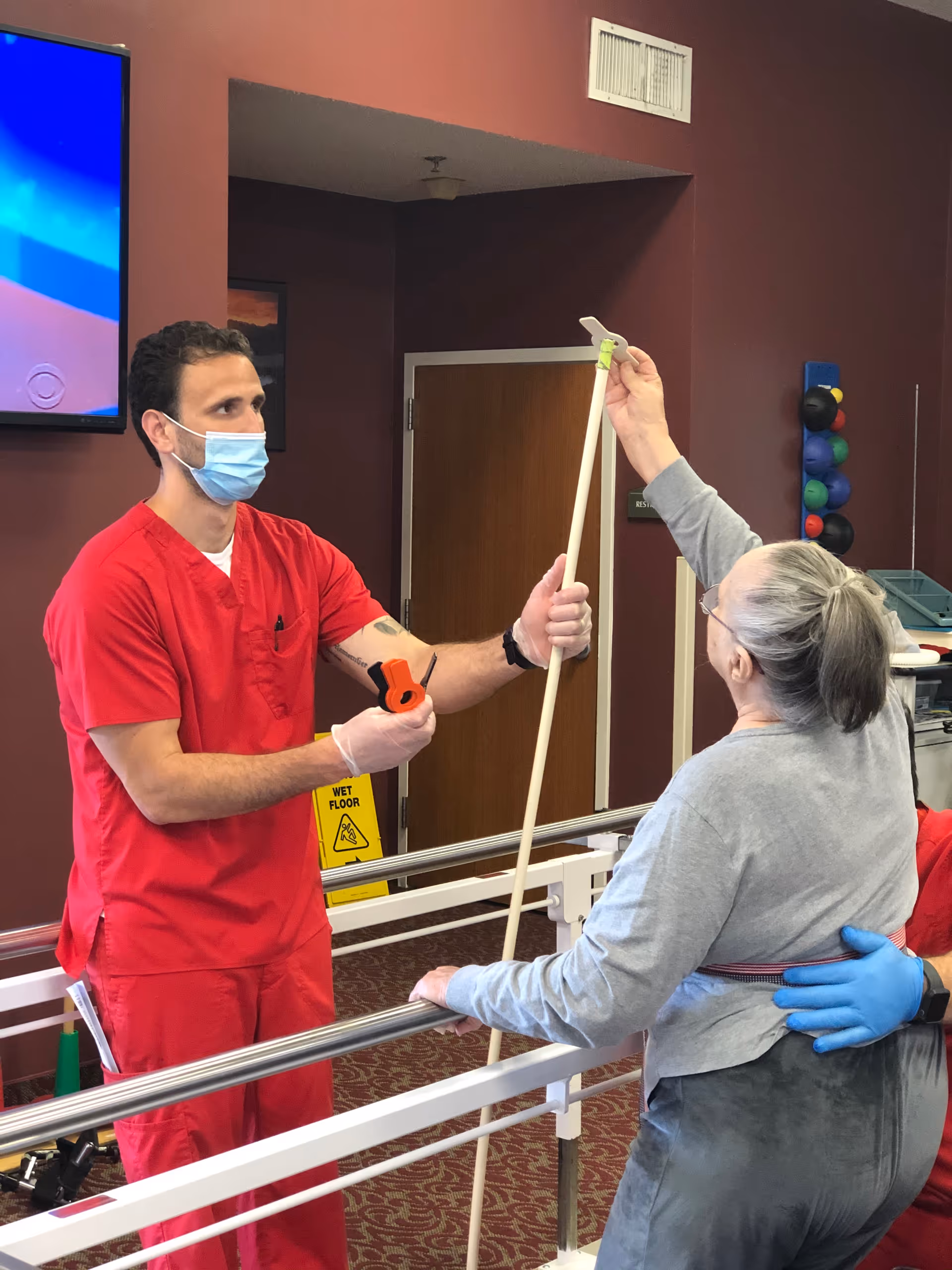 A healthcare worker in red scrubs and a face mask assists an elderly woman with gray hair in a ponytail during a physical therapy session. The woman is holding a long stick and standing between parallel bars, while the worker supports her and holds a measuring tape. The room has maroon walls, a wooden door, and exercise balls mounted on the wall in the background.