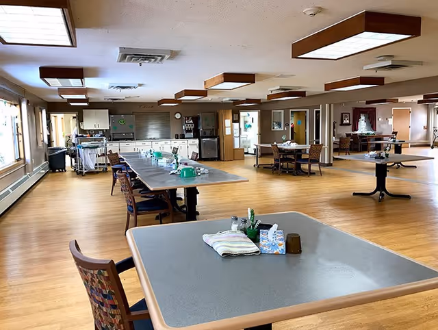 A spacious dining room with multiple tables and chairs arranged on a wooden floor. The tables have various items on them including napkins, tissue boxes, and condiments. The room has large windows on one side allowing natural light to enter, and the ceiling features rectangular light fixtures. In the background, there is a kitchen area with cabinets and appliances.