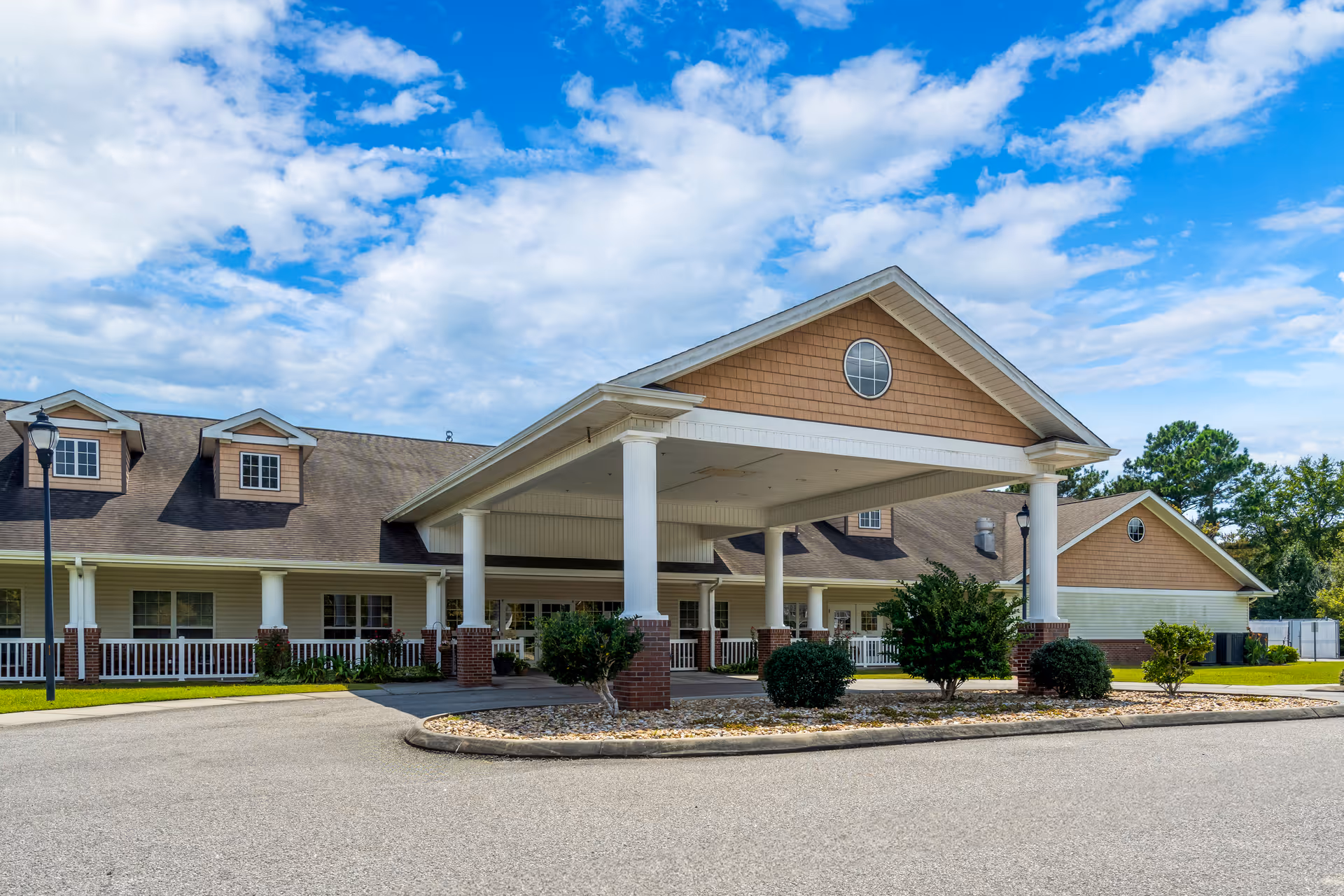 Exterior view of a single-story rehabilitation and healthcare center building with a covered entrance supported by white columns. The building has a brown shingled roof with dormer windows and a circular window on the front gable. There are bushes and small trees around the entrance area under a partly cloudy blue sky.