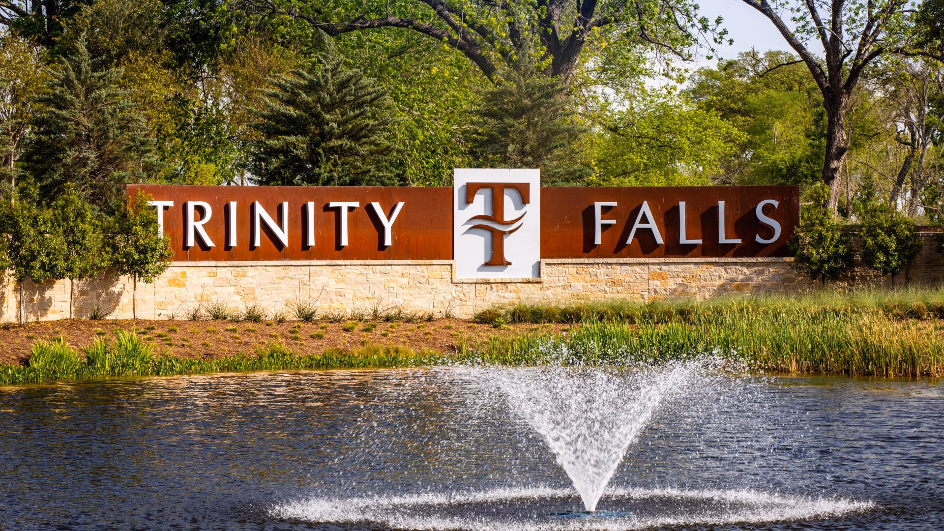 Stone and metal 'Trinity Falls' community entrance sign by a pond with a fountain and trees behind it.
