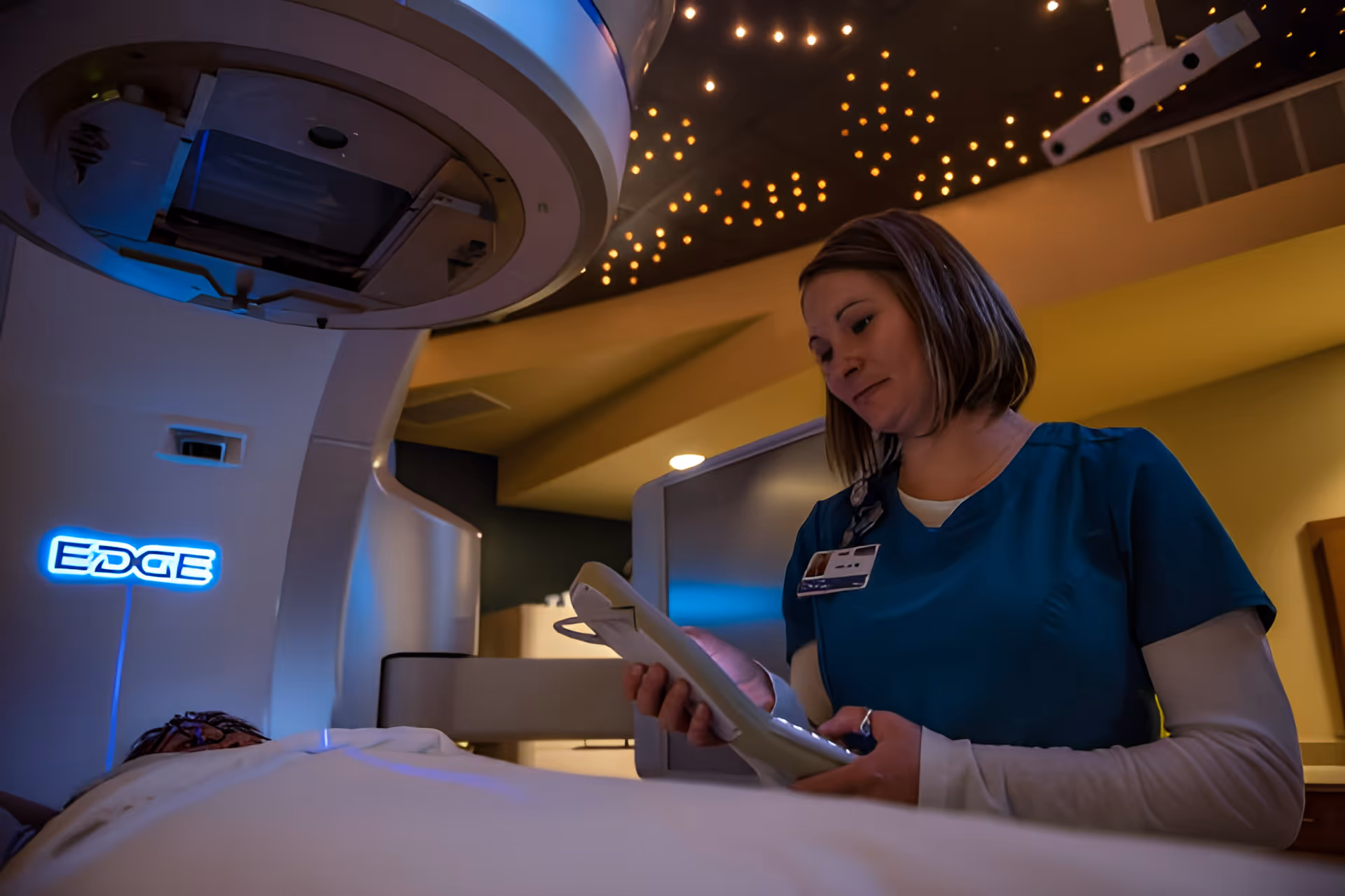 A medical technician in teal scrubs uses a handheld control next to a large 'EDGE' treatment machine in a dimly lit procedure room.