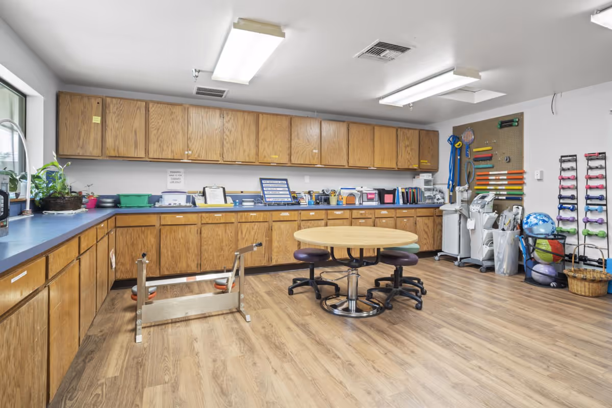 A spacious room with wooden cabinets lining two walls, a round table with four stools in the center, and various exercise and therapy equipment including colorful dumbbells, exercise balls, and resistance bands organized on racks and a pegboard. The room has wood flooring and bright overhead lighting.