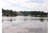 A calm lake with reflections of the sky and surrounding trees. In the background, there are green trees and a few buildings partially visible through the foliage under a partly cloudy sky.