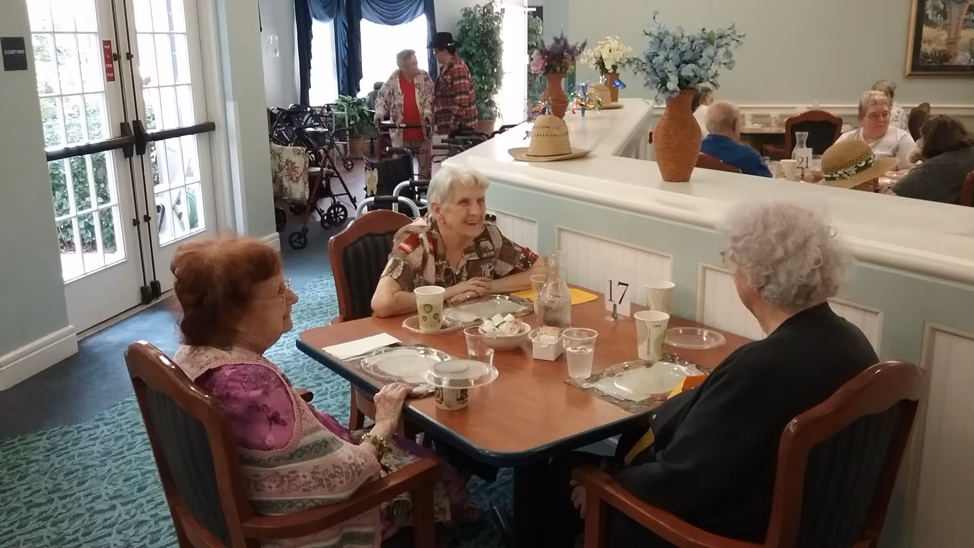 Three elderly women sitting around a dining table in a senior living facility, with plates, cups, and a water pitcher on the table. In the background, other elderly people are seated at tables, and two women are standing and talking near a window with walkers nearby. The room is decorated with vases of flowers and has a light, welcoming atmosphere.