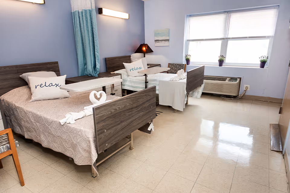 A bright room with two hospital-style beds, each with a pillow that says 'relax' and a towel folded into a swan shape on the foot of the beds. The room has light blue walls, a window with blinds, an air conditioning unit below the window, and a wooden chair in the corner.