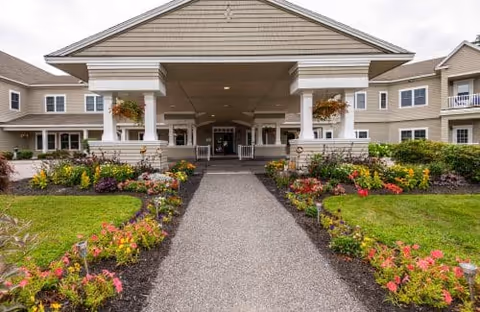 Front exterior view of a senior living facility with a covered entrance, stone pillars, and a pathway lined with colorful flowers and green lawns on both sides.