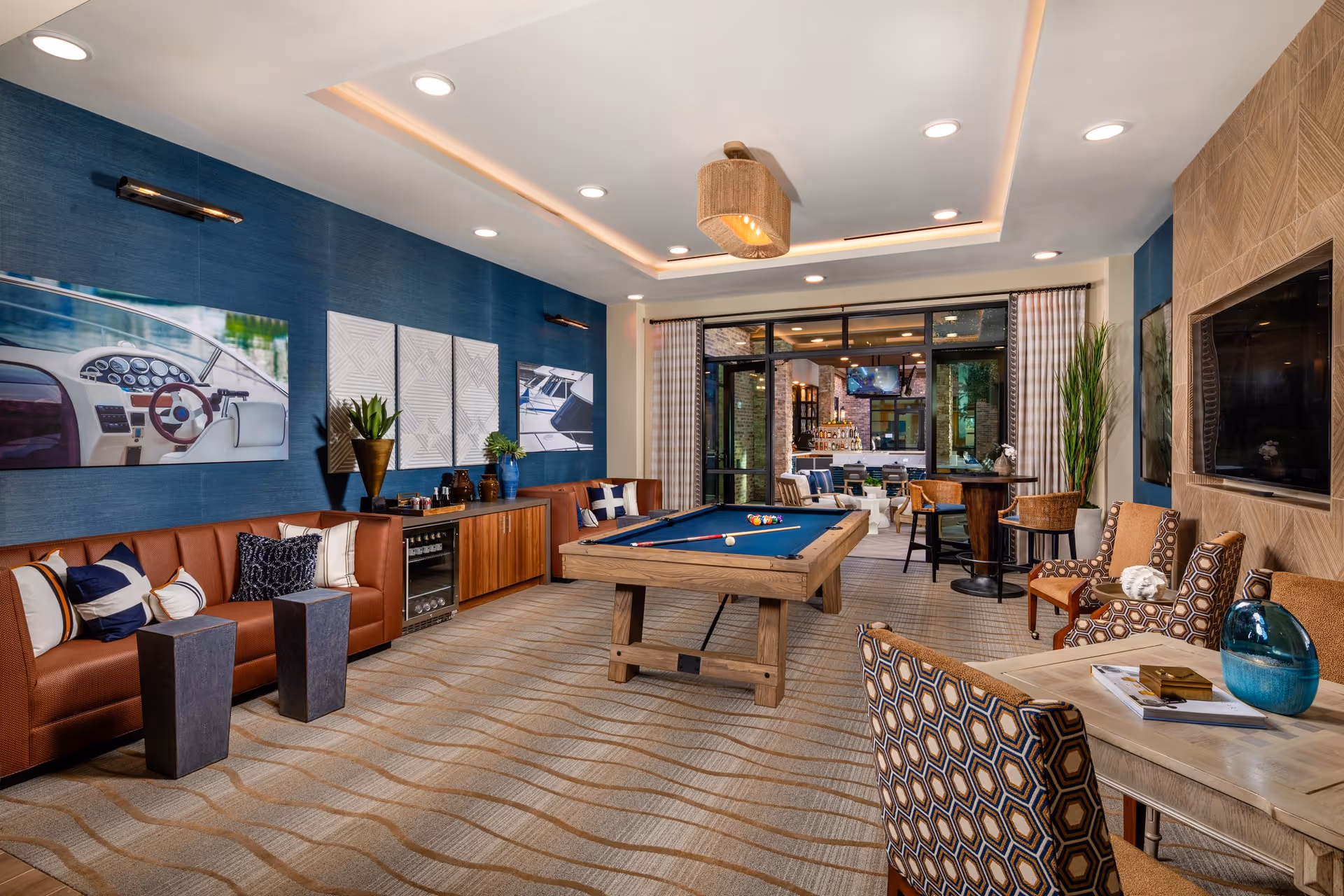 A stylish and modern common area featuring a pool table in the center, surrounded by comfortable seating including leather couches, patterned chairs, and small tables. The room has blue accent walls with nautical-themed artwork, recessed ceiling lights, and large windows leading to an adjacent room with more seating and a bar area.