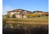 A brick senior living facility with multiple wings sits beside a pond and grassy grounds.