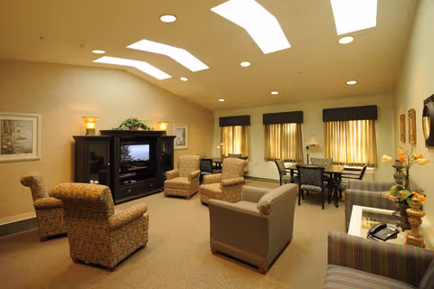 A spacious senior living common area with multiple upholstered armchairs and sofas arranged around a large entertainment center with a television. The room has beige walls, carpeted floor, and several windows with curtains. There is a table with chairs near the windows and decorative plants and lamps placed around the room.