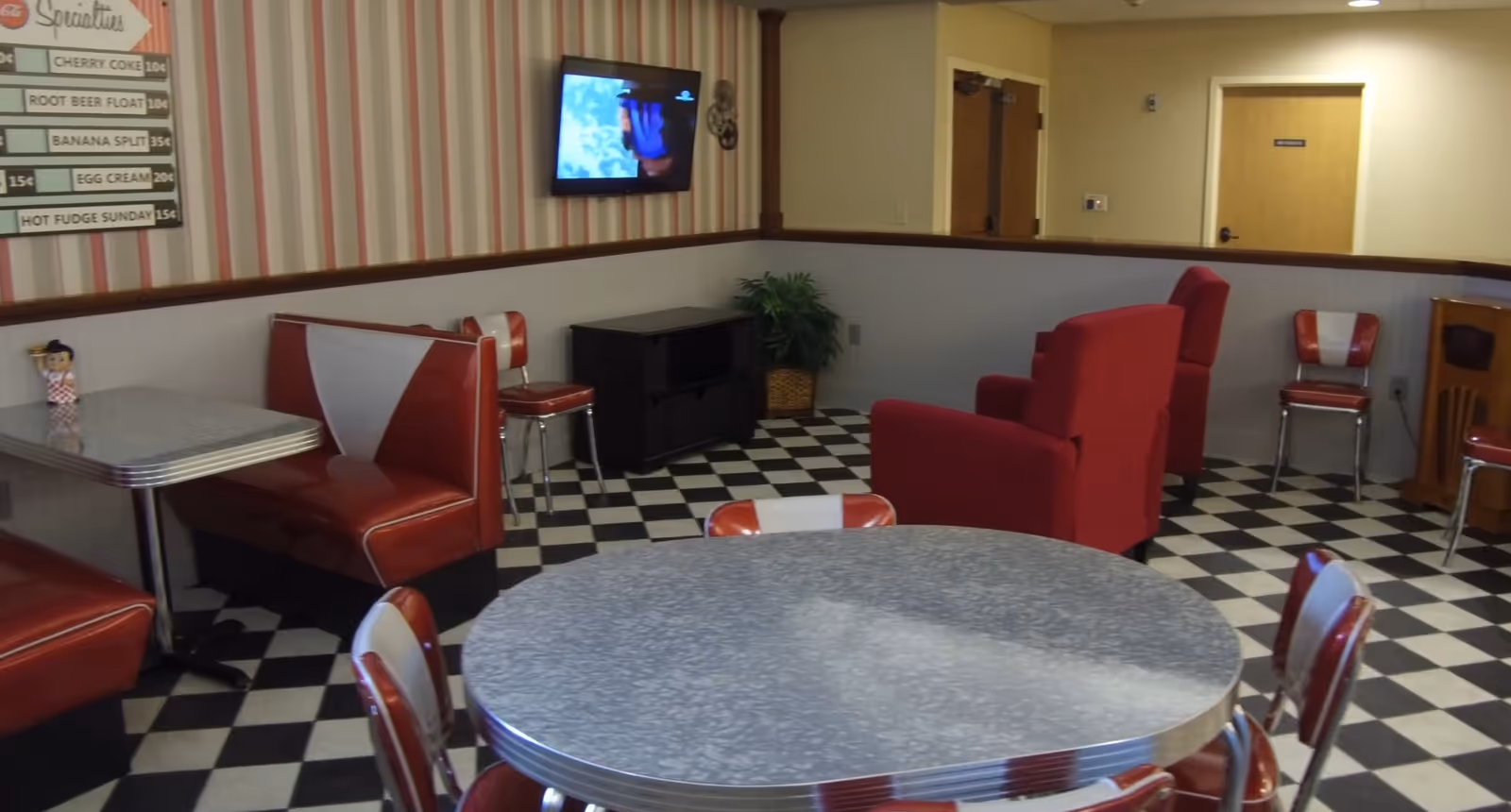 Retro-style common room with black-and-white checkered floor, red diner booths and chairs, round tables, and a wall-mounted TV.