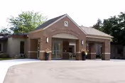 Front entrance of a single-story brick senior care building with a covered porte-cochere, glass double doors, and potted plants.