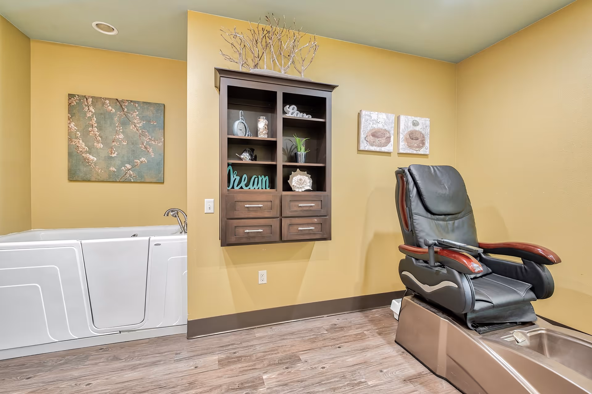 A room with a walk-in bathtub on the left and a black pedicure spa chair on the right. The walls are painted yellow, and there is a wooden wall cabinet with decorative items and artwork hanging on the walls.