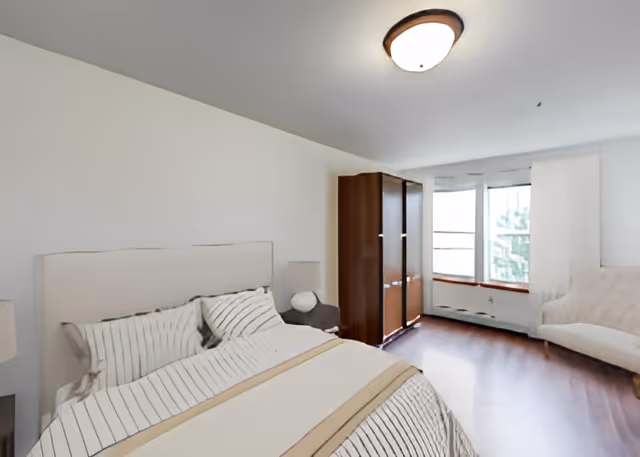 A bright bedroom with a large bed featuring striped bedding, a wooden wardrobe, a cushioned chair, and a window with white curtains letting in natural light.