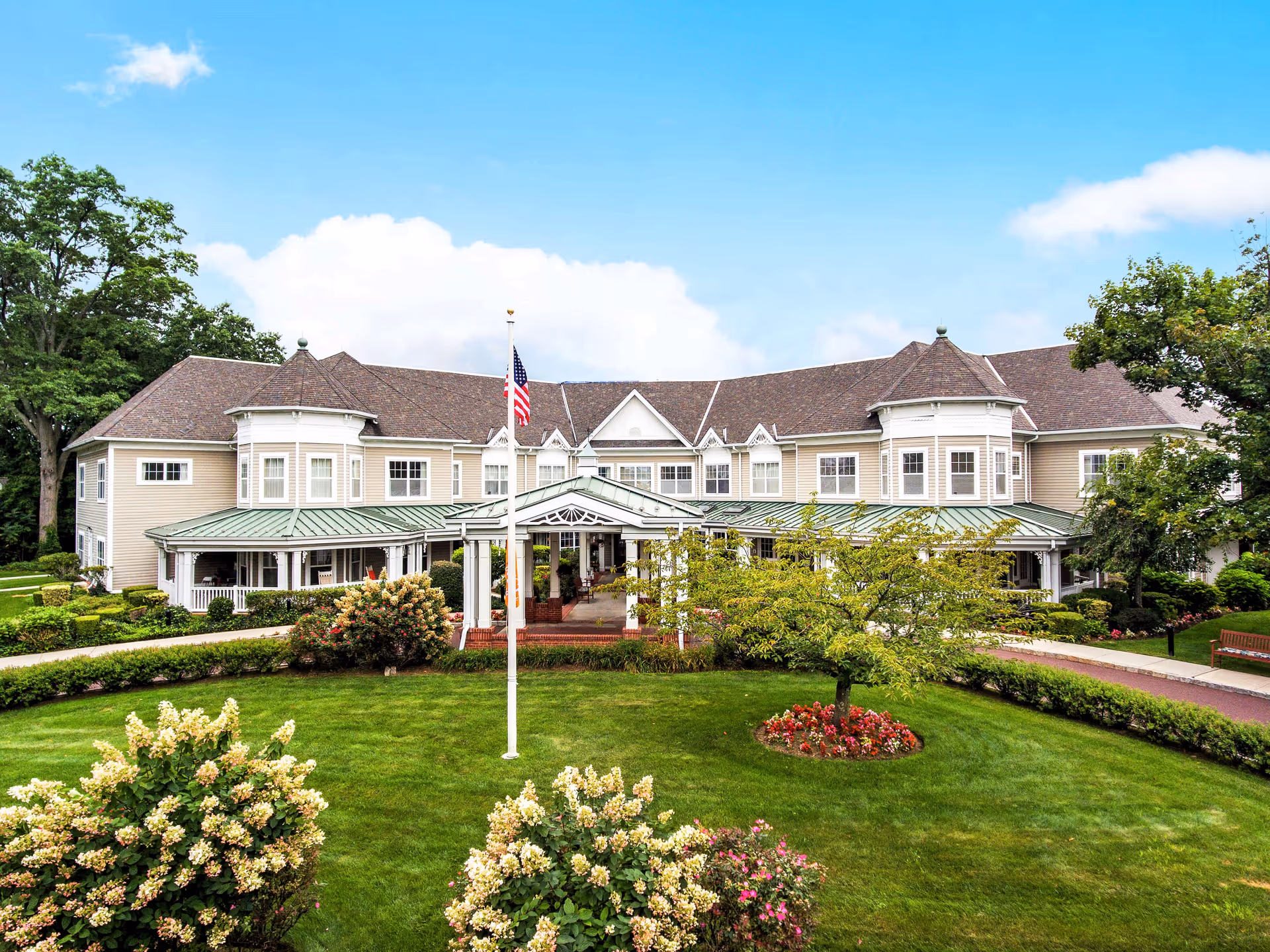 Front exterior view of a large senior living facility building with beige siding, multiple windows, and a green metal roof over the entrance. The building is surrounded by well-maintained green lawns, flower beds, bushes, and trees. An American flag is displayed on a flagpole in front of the entrance under a covered porch.