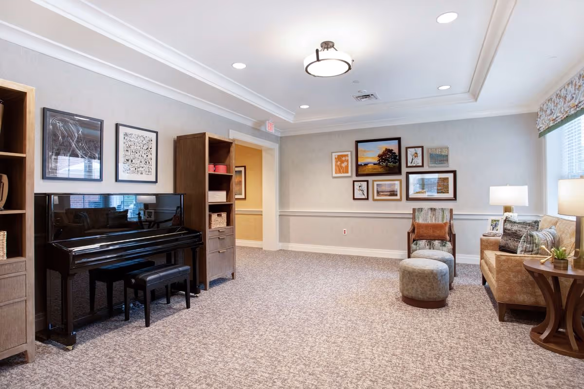 Well-lit common room with a black upright piano, bookshelves, framed artwork, and a seating area with chairs and a sofa.