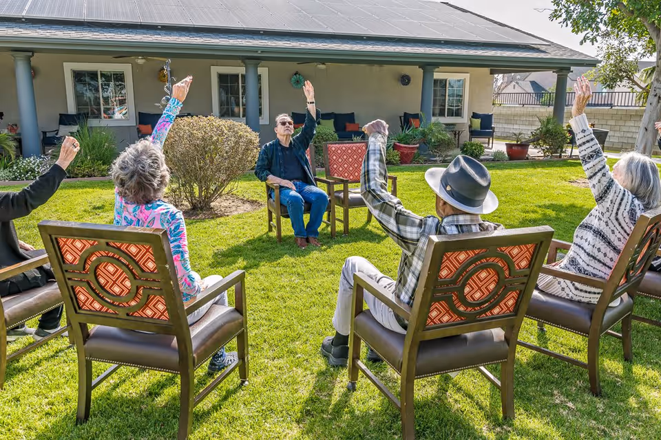 A group of elderly people sitting in a circle on outdoor chairs on a grassy lawn, participating in a seated exercise or stretching activity with their arms raised. They are in front of a building with a covered porch and several windows.