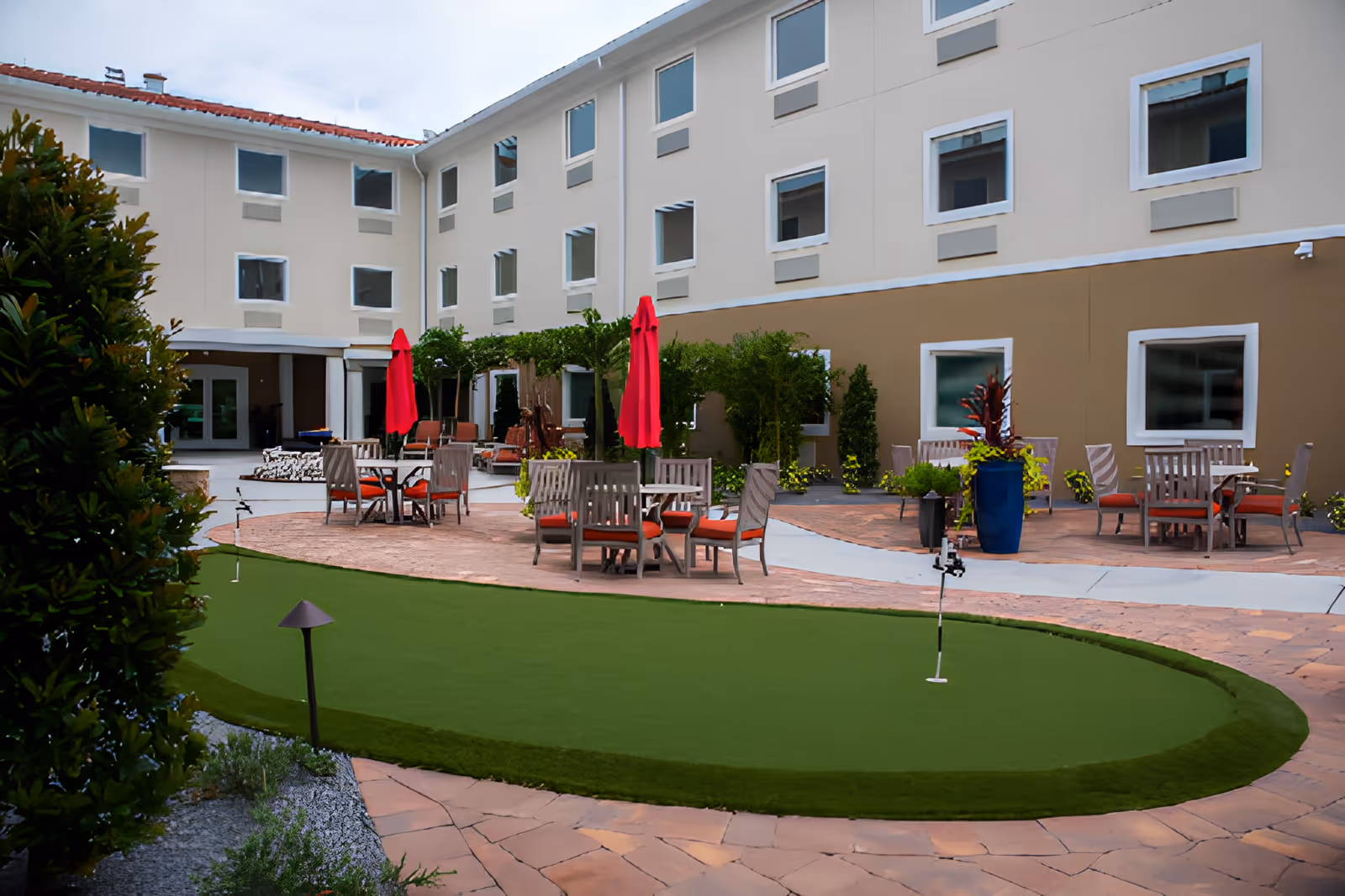 Outdoor courtyard with a small putting green, patio tables and red umbrellas in front of a three-story building.