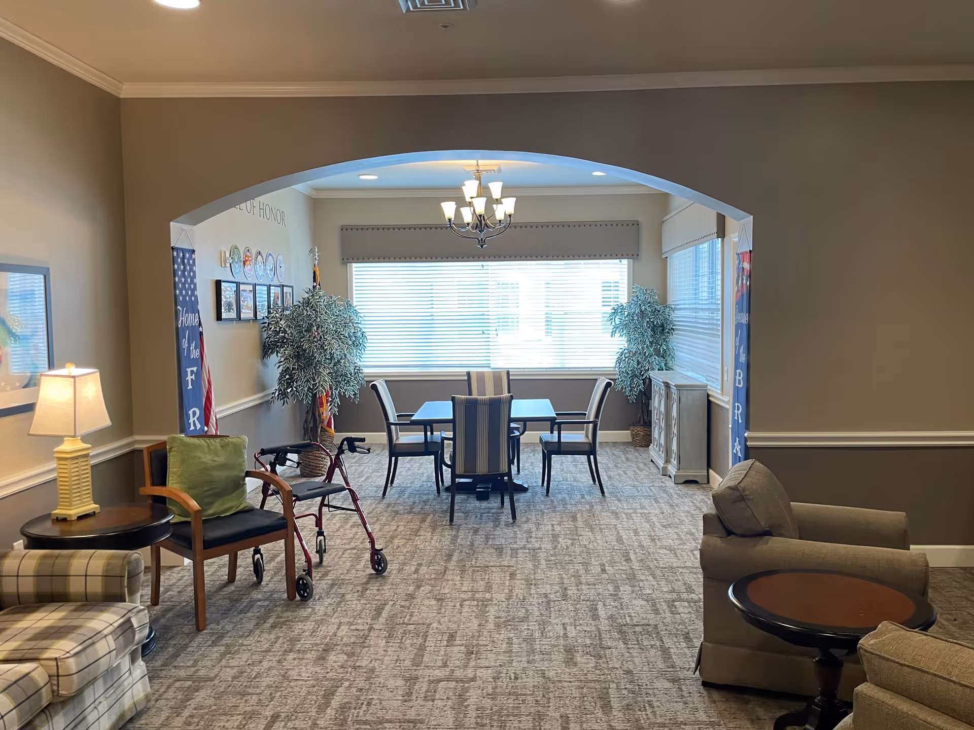 A cozy interior common area in a senior living facility with a carpeted floor, an archway leading to a dining area with a table and four chairs. The room has two large windows with blinds, two potted plants, and a chandelier. In the foreground, there are armchairs, a side table with a lamp, and a walker next to a chair with a green cushion. The walls are decorated with framed pictures and banners.