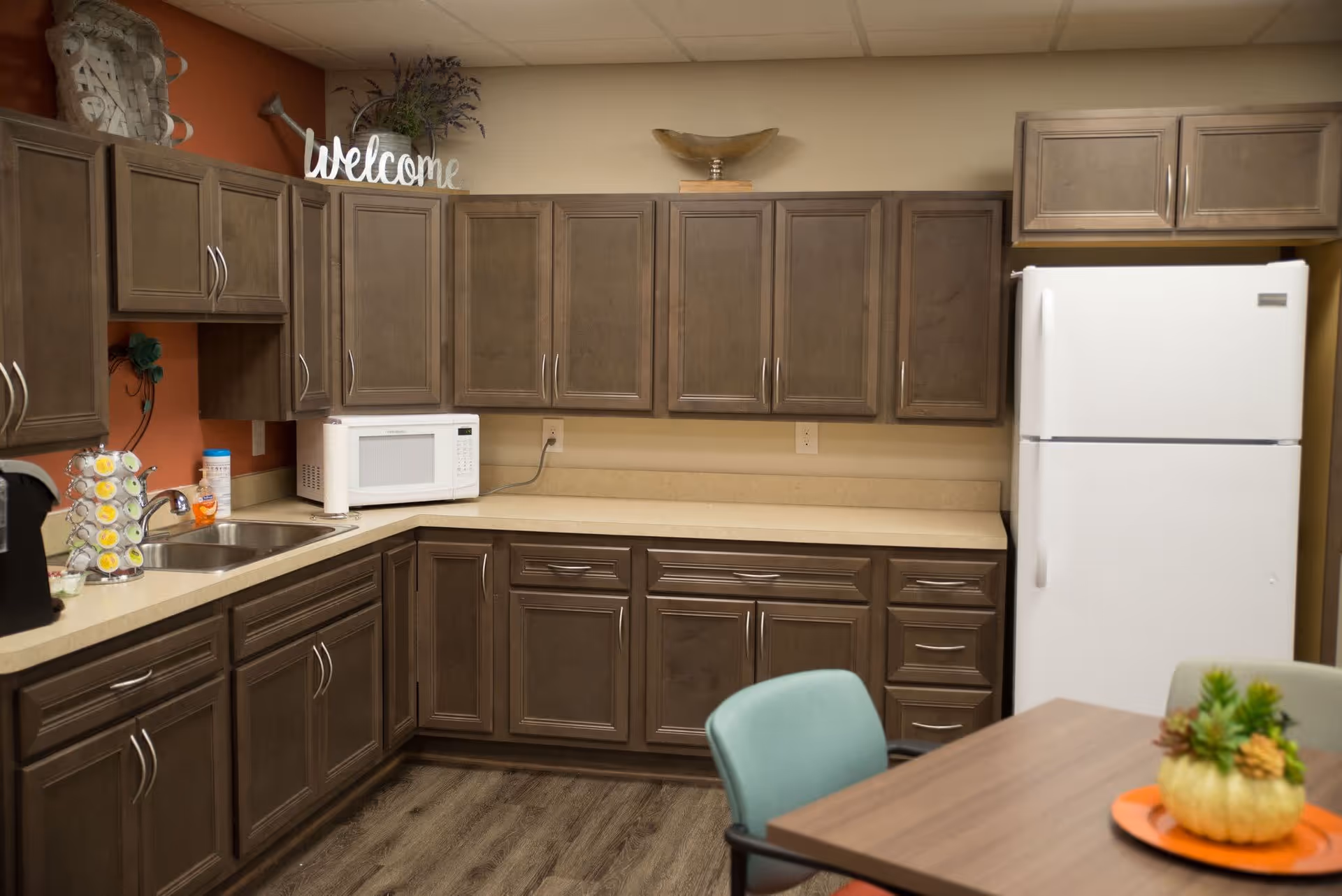 A kitchen area with brown cabinets, a white microwave, a white refrigerator, a double sink, and a coffee pod holder. There is a wooden table with a small decorative plant on an orange tray and a green chair. The wall has a decorative 'welcome' sign and some plants on top of the cabinets.
