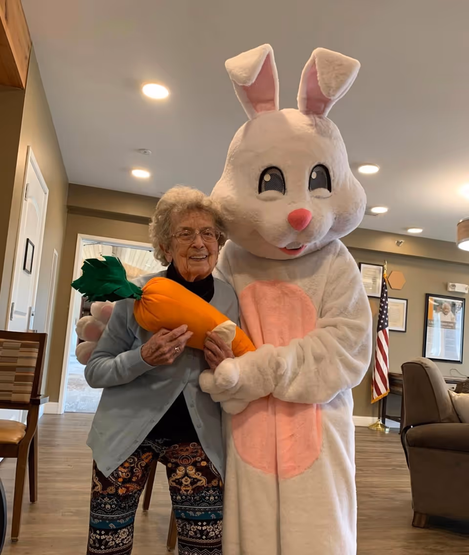 An elderly woman standing indoors next to a person dressed in a large white and pink bunny costume holding a large plush carrot. The woman is smiling and wearing glasses, a light blue cardigan, and patterned pants. The room has wooden flooring, chairs, an American flag, and framed pictures on the wall.