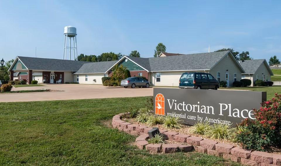 Front view of the Victorian Place senior residential care building with its sign, parking area, and a water tower in the background.