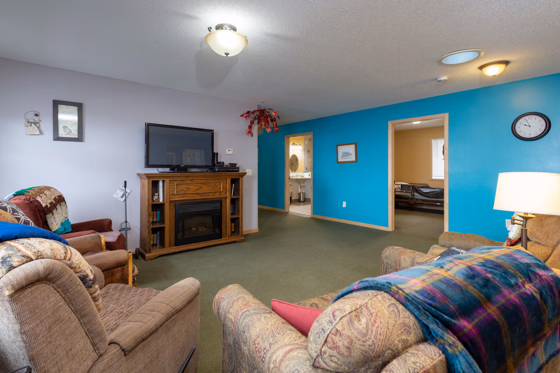 A cozy living room with multiple upholstered armchairs and a sofa, a wooden TV stand with a flat-screen television mounted above an electric fireplace, and a blue accent wall with two doorways leading to a bathroom and a bedroom. The room has green carpet flooring, a ceiling light fixture, and a wall clock above a table lamp.