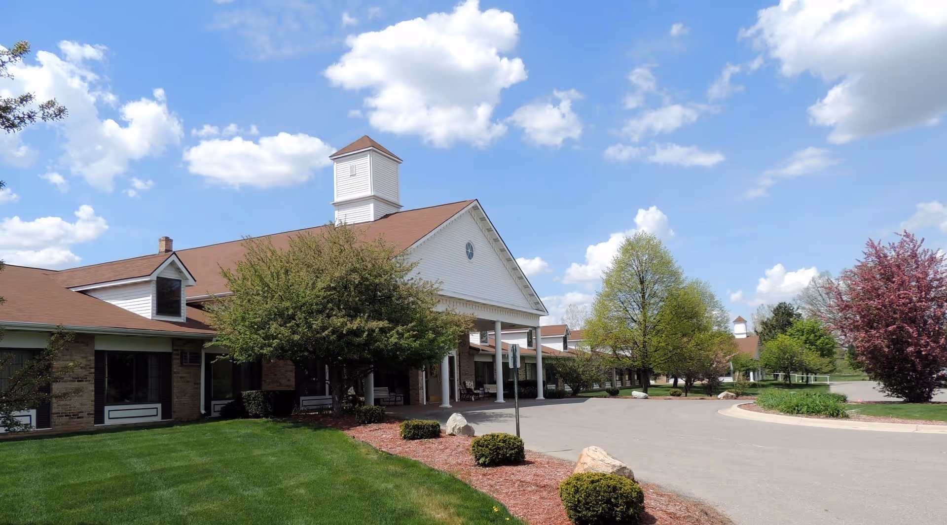 Exterior view of a single-story retirement center building with a peaked roof and a small tower. The building is surrounded by well-maintained green lawns, bushes, and trees under a partly cloudy blue sky.
