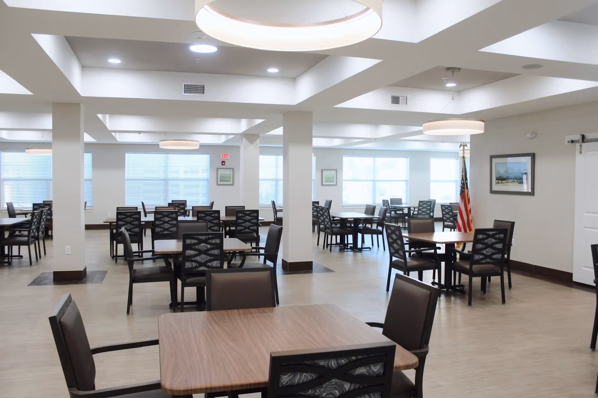 A spacious, well-lit dining room with multiple square wooden tables and black chairs arranged neatly. Large windows allow natural light to fill the room. The walls are light-colored, and there are modern circular ceiling lights. An American flag stands in one corner near a framed picture on the wall.