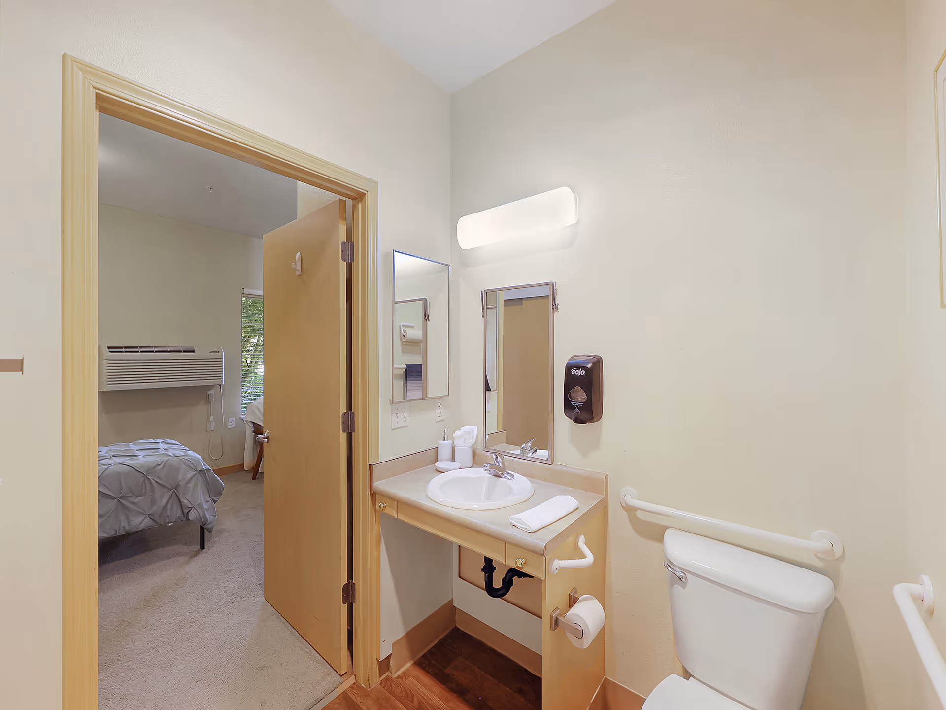 View of a small bathroom with a white toilet, a sink with a mirror above it, a soap dispenser on the wall, and grab bars for accessibility. The bathroom door is open, showing a bedroom with a bed and a window with blinds.