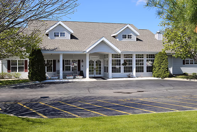 Front exterior view of a single-story building with a gabled roof, white siding, multiple windows, and a covered entrance. There are trees and shrubs around the building, a paved parking lot with yellow-striped parking spaces, and a green lawn in the foreground under a clear blue sky.