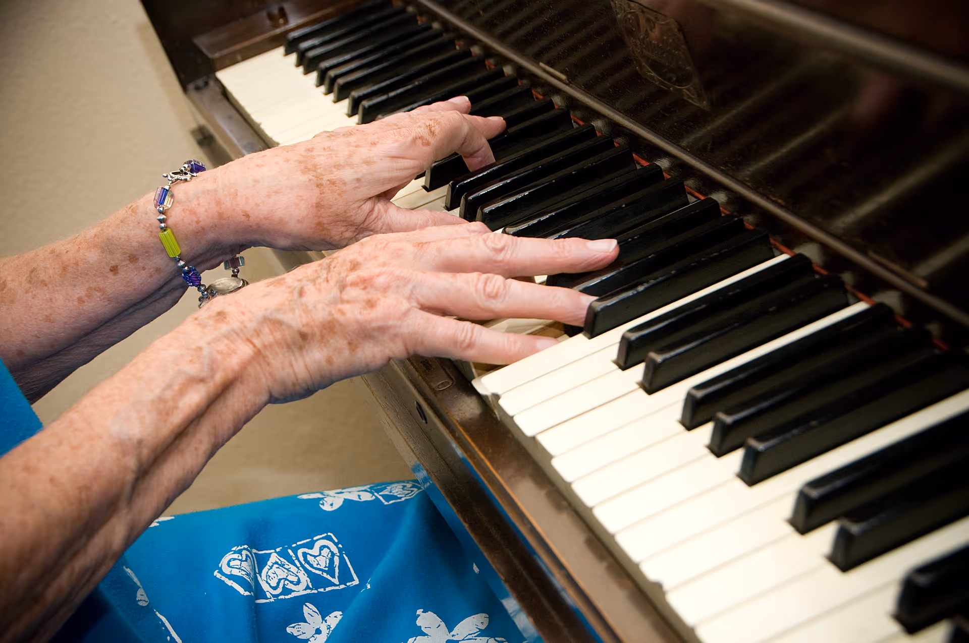 Close-up of elderly hands playing the keys of a piano, with a colorful bracelet on one wrist and a blue garment with white patterns visible.