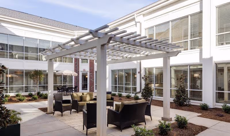 Outdoor courtyard area with a white pergola providing partial shade over a seating arrangement consisting of cushioned chairs and a sofa. The courtyard is surrounded by a two-story building with large windows and some landscaping including small bushes and trees.