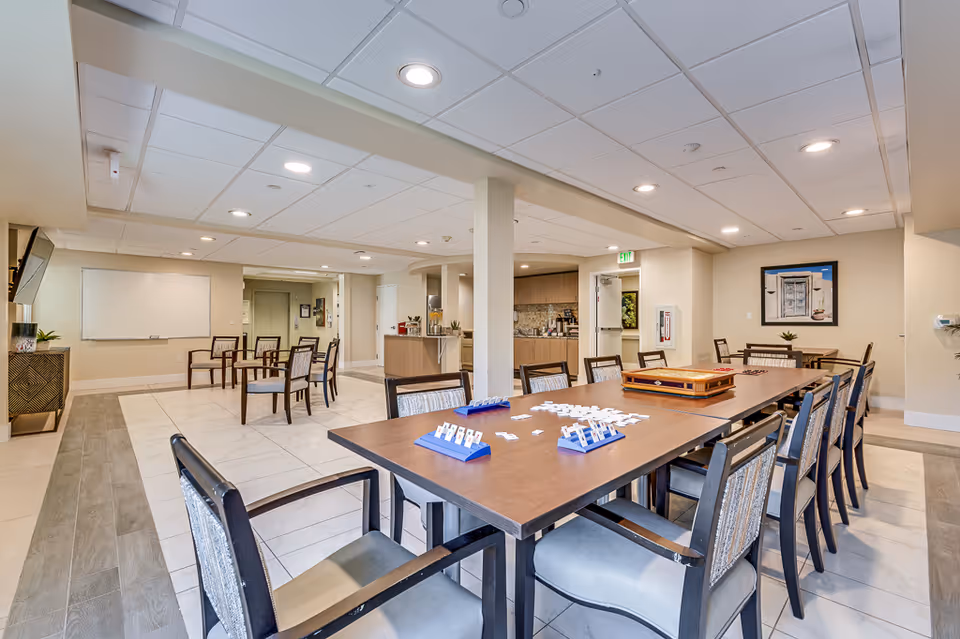Bright communal activity/dining room with a long table and chairs set up for games and a kitchenette and seating area in the background.