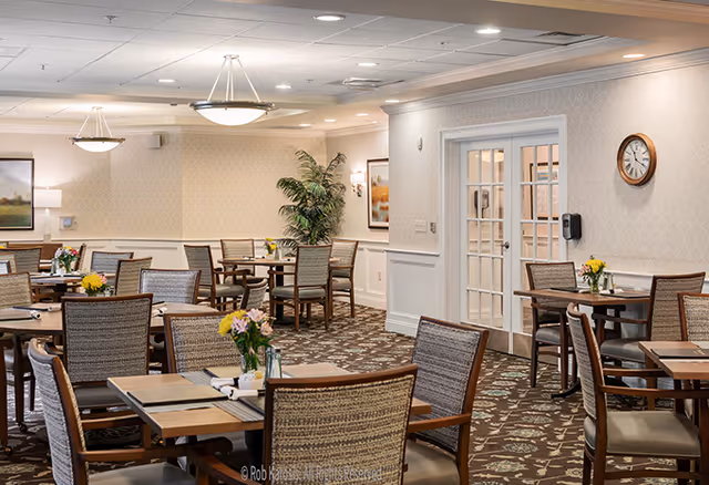 A well-lit senior dining room with multiple wooden tables and cushioned chairs arranged neatly. Each table has placemats and small flower arrangements. The room features patterned carpet, light-colored walls with framed artwork, a large potted plant, and ceiling light fixtures. A clock is mounted on the wall near a set of double glass doors.