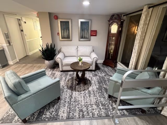 A cozy seating area in a senior living facility with a white loveseat, two light blue armchairs, a round wooden coffee table with a small plant, a large patterned rug, a tall grandfather clock, two framed artworks on the wall, and a large potted plant near a hallway.