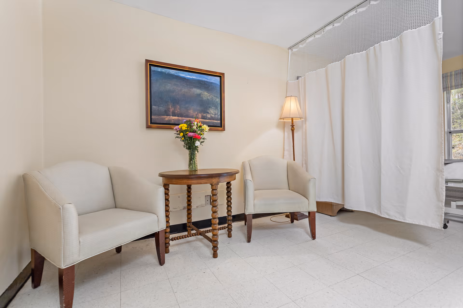 A small seating area with two beige armchairs and a round wooden table with a vase of colorful flowers between them. A floor lamp stands next to one chair, and a large white privacy curtain is partially drawn in the background. A framed landscape painting hangs on the wall above the table.