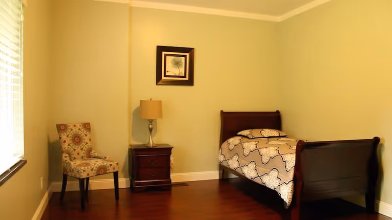 A simple bedroom with light green walls, a wooden single bed with patterned bedding, a wooden nightstand with a lamp, a framed picture on the wall, and a patterned upholstered chair near a window with blinds.