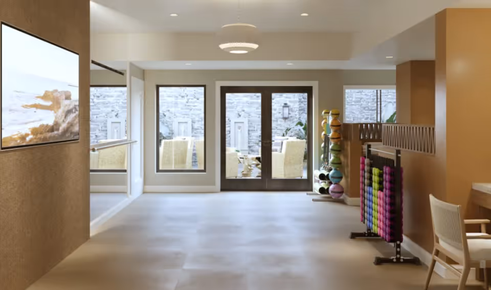 Bright interior common area with large windows and double doors to a patio, TV on the left wall and racks of exercise balls and weights on the right.