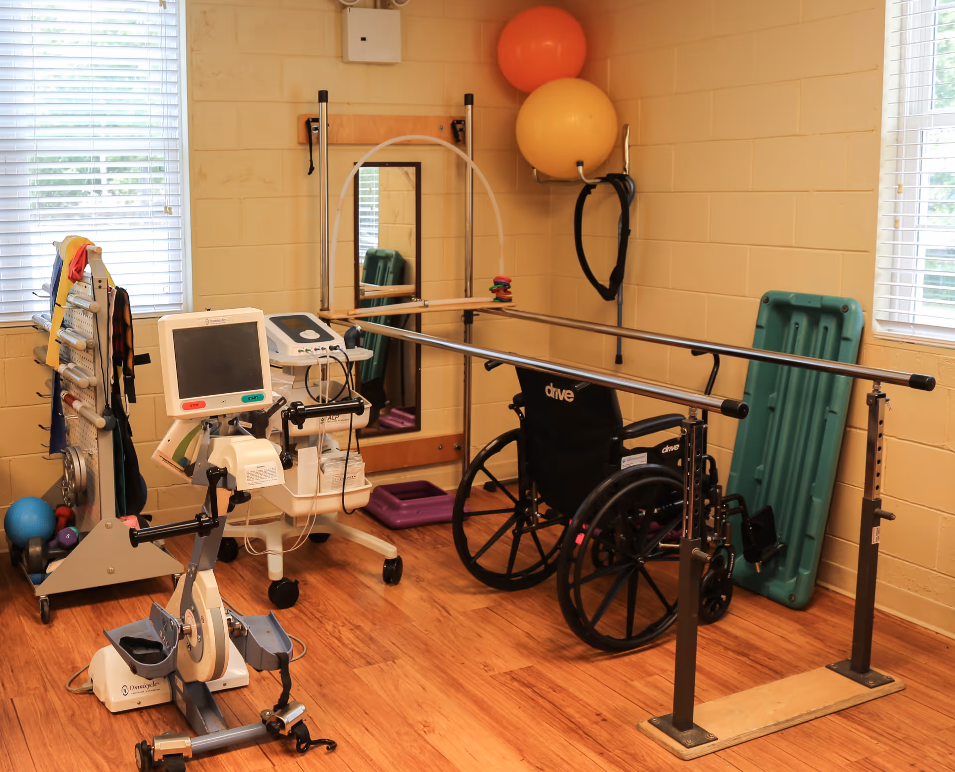 A rehabilitation room with parallel bars for walking exercises, a wheelchair, exercise balls mounted on the wall, a stationary exercise bike with a monitor, resistance bands hanging on a rack, and a mirror on the wall. The room has wooden flooring and two windows with blinds.