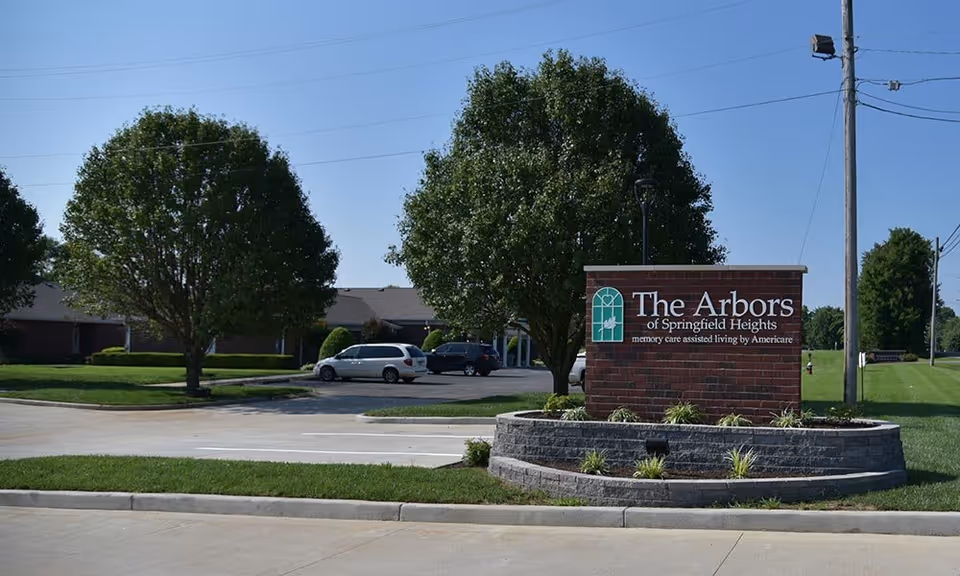 A landscaped entry sign reading "The Arbors of Springfield Heights" in front of a low brick building, parking lot, and trees under a clear sky.
