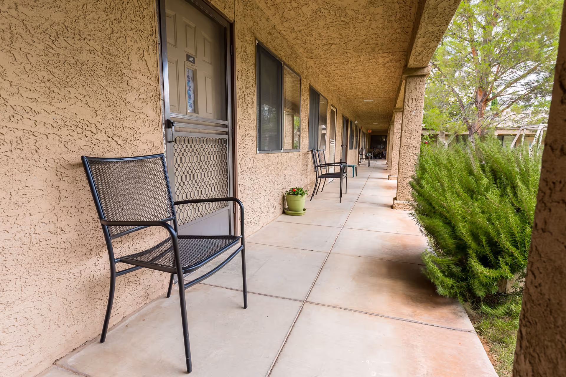 A covered outdoor walkway at Verde Valley Assisted Living with beige textured walls and several black metal chairs placed along the corridor. There are doors and windows along the wall, a small green potted plant on the ground, and green bushes on the right side of the walkway.