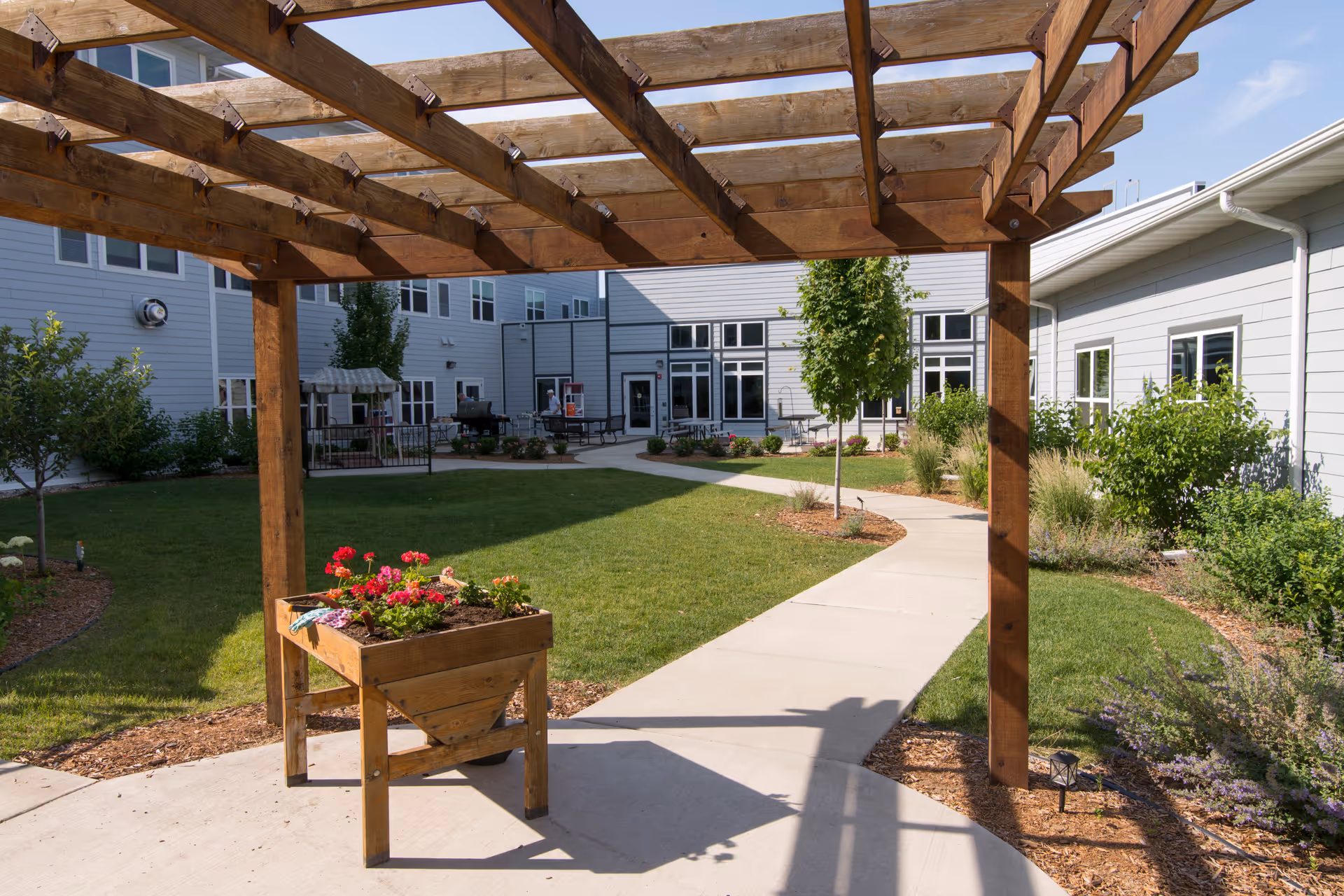 Outdoor courtyard area at Sunrise Retirement Community featuring a wooden pergola, a raised planter box with red and pink flowers, a concrete walkway, green grass, small trees, and shrubs surrounding the space with the facility building in the background.