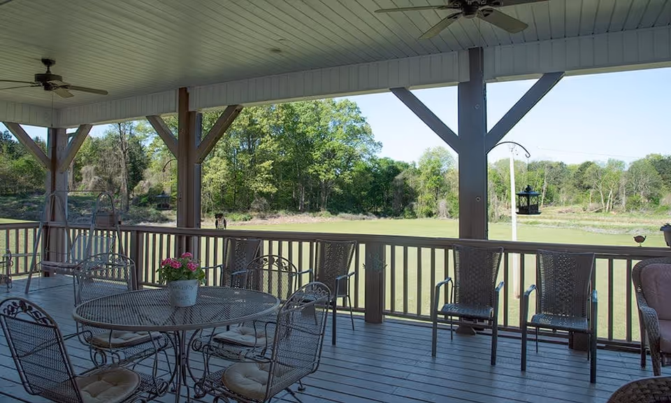 Covered outdoor patio area with metal table and chairs, some with cushions, overlooking a grassy field and trees in the background under a clear sky.