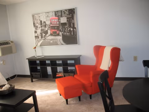 A living room with a red wingback chair and matching ottoman, a black shelving unit, wall art of a red double-decker bus, and a small table with an air conditioner on the left wall.