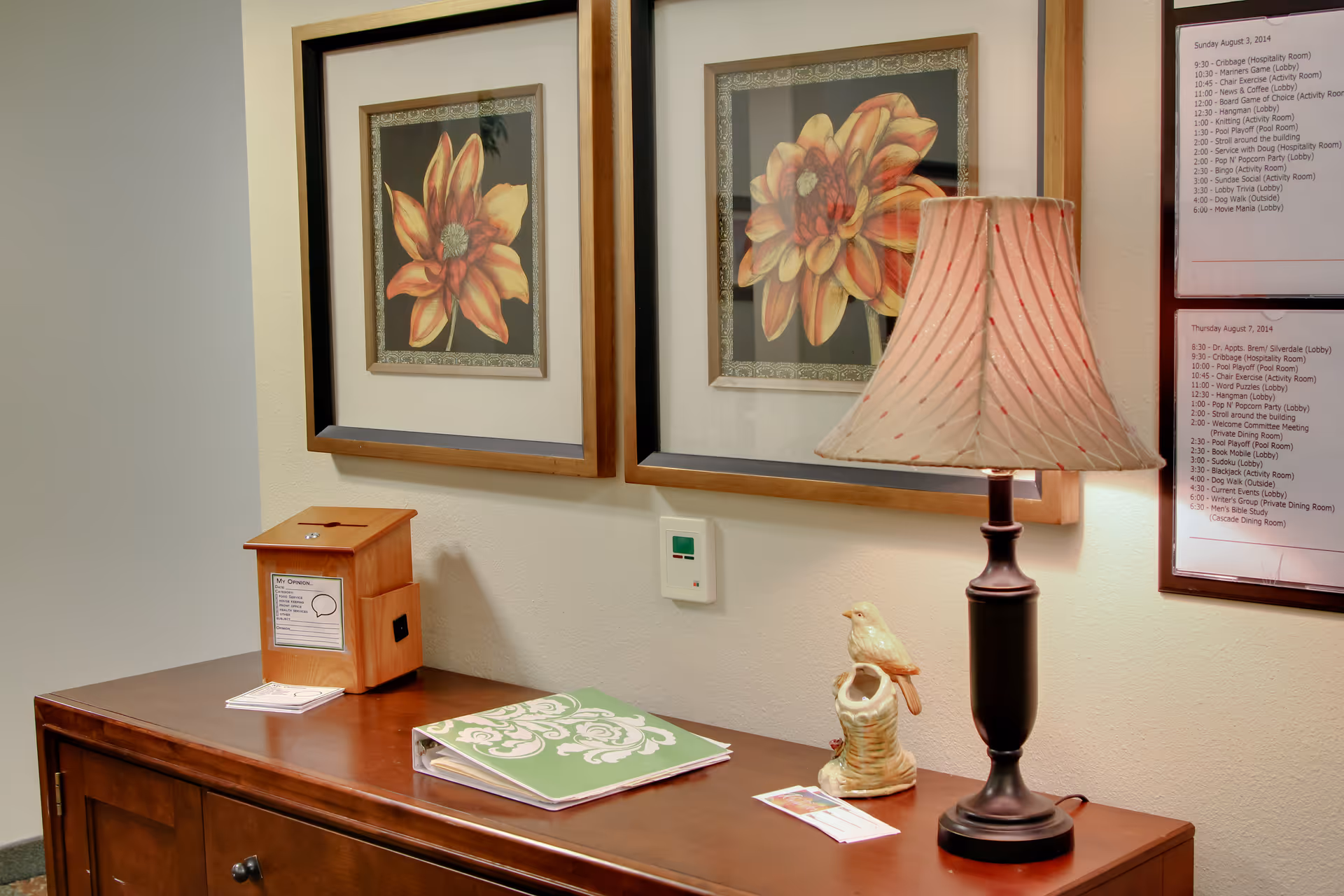 A wooden sideboard with a decorative lamp, a ceramic bird figurine, a green patterned binder, and a wooden suggestion box. Above the sideboard are two framed floral artworks and a thermostat on the wall. To the right, two framed schedules are partially visible on the wall.