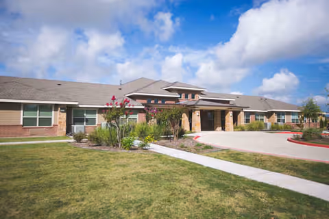 Single-story brick and stone building with a sloped roof under a partly cloudy blue sky, surrounded by a green lawn and landscaped bushes, with a curved driveway leading to the entrance.