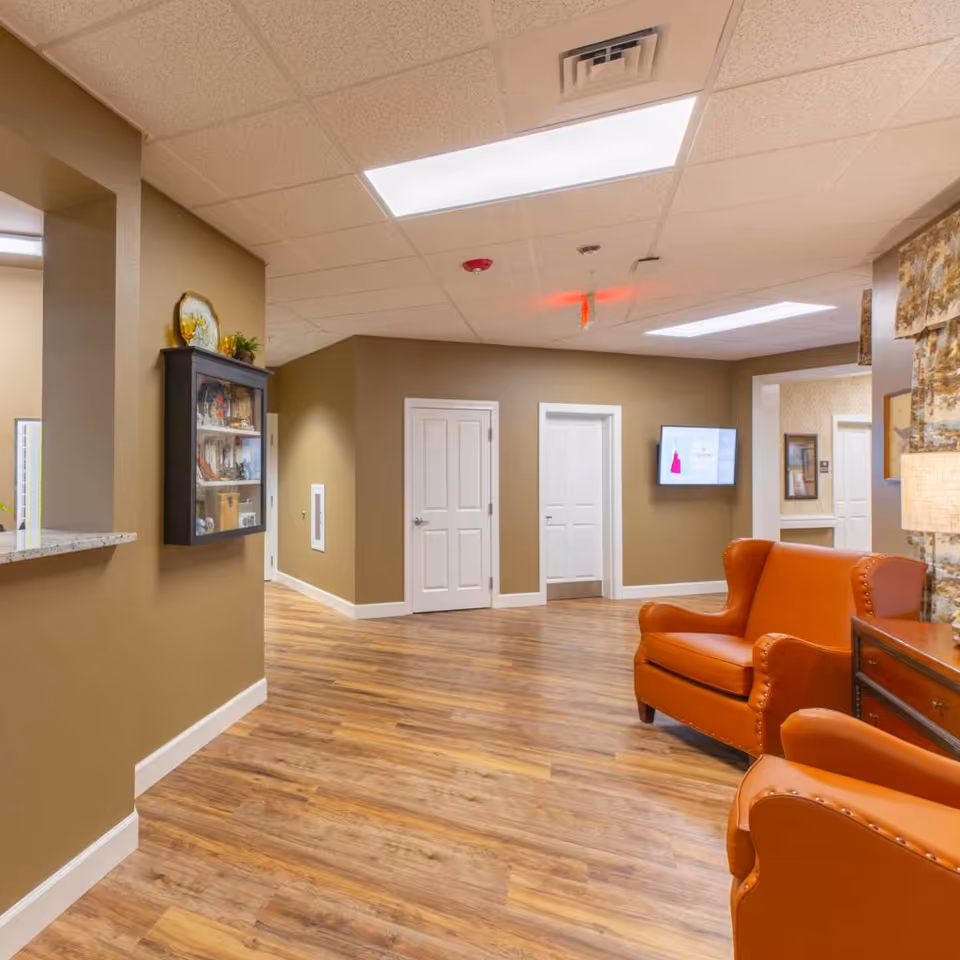 Interior view of a senior living facility hallway with wood flooring, beige walls, and white doors. There are two orange leather armchairs on the right side next to a wooden side table with a lamp. A wall-mounted display case with decorative items is on the left wall, and a flat screen TV is mounted on the far wall. The ceiling has recessed lighting panels.