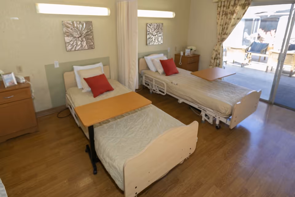 Two adjustable hospital beds with overbed tables and bedside cabinets in a bright shared patient room with a sliding glass door to a patio.