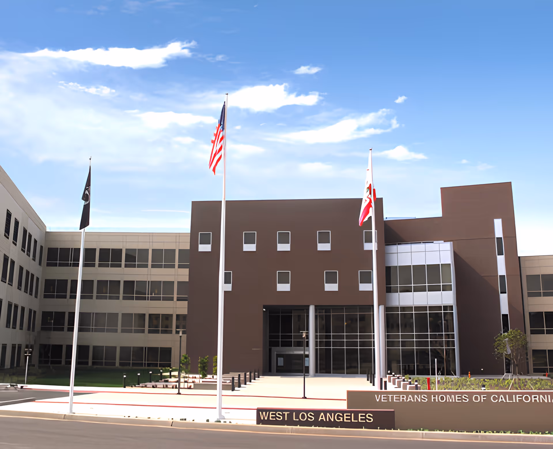 Front exterior view of the Veterans Home of California - West Los Angeles building with three flagpoles displaying the American flag, a black flag, and the California state flag. The building is modern with multiple windows and a clear blue sky above.